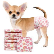 Fawn Chihuahua wearing Dono disposable pull-up female dog diaper in pink paw-print pattern, side view facing camera, with stack of folded diapers showing yellow wetness indicator lines, on white background