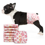 Black-and-white Boston Terrier puppy wearing Dono disposable pull-up female dog diaper in pink paw-print pattern, side view, with stack of folded diapers showing yellow wetness indicator lines, on white background
