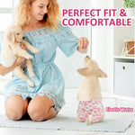 Young woman in blue dress kneeling indoors holding Golden Retriever puppy while second puppy wearing Dono disposable pull-up female dog diaper in pink paw-print pattern stands on hind legs, 